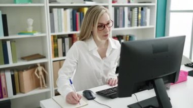 Young blonde woman student using computer writing on notebook at library university