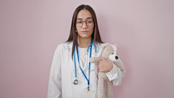 年轻美丽的惊慌失措的女医生抱着泰迪熊 默默无闻地站在孤立的粉色背景之上 — 图库视频影像