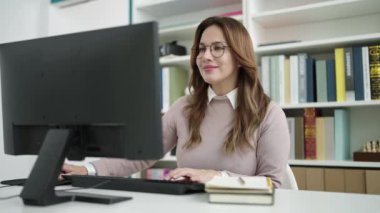 Young beautiful hispanic woman student using computer writing on notebook at library university