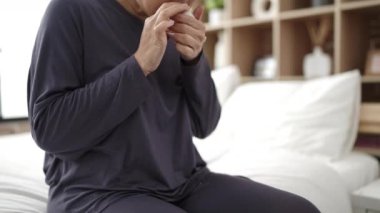 Young chinese woman applying skin lotion treatment sitting on bed at bedroom