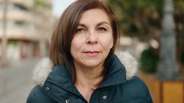 Orta yaşlı kadın gülümsüyor. Kollarını kavuşturup caddede duruyor.