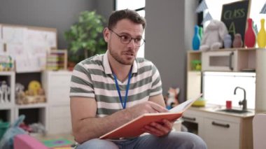 Young caucasian man working as teacher looking at notebook explaining at kindergarten