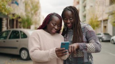Two african american women smiling confident using smartphone at street