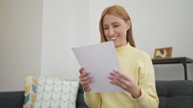 Young blonde woman reading document sitting on sofa at home