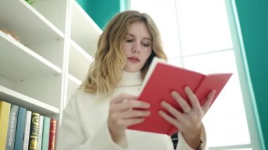 Young blonde woman student reading book standing at library university