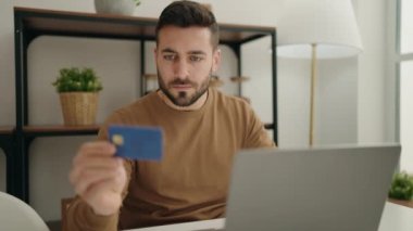 Young hispanic man using laptop and credit card sitting on table at home