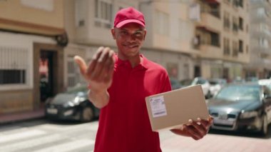 Young latin man delivery worker holding package doig coming gesture with hand at street