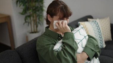 Young caucasian woman crying sitting on the sofa hugging pillow at home