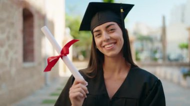 Young hispanic woman wearing graduated uniform holding diploma at university