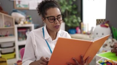 African american woman preschool teacher reading book sitting on chair at kindergarten