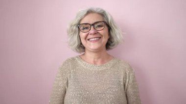 Gri saçlı, kendine güvenen, soyutlanmış pembe arka planda duran orta yaşlı bir kadın.