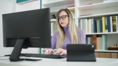 Young blonde woman student using computer and touchpad studying at library university