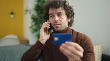 Young hispanic man talking on smartphone holding credit card sitting on sofa at home