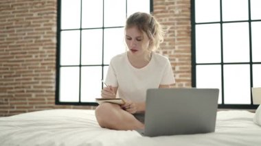 Young blonde woman using laptop writing on notebook sitting on bed at bedroom