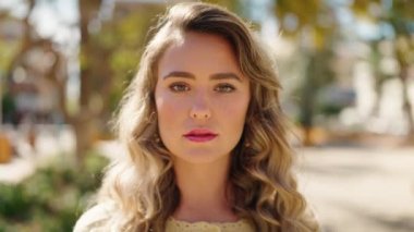 Genç sarışın kadın gülümsüyor. Parkta kollarını kavuşturmuş duruyor.