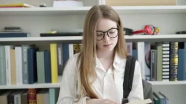 Young blonde woman student reading book doing silence gesture at university classroom