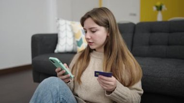 Akıllı telefon ve kredi kartı kullanan genç sarışın kadın evde yerde oturuyor.