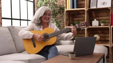 Middle age grey-haired woman smiling confident having online guitar class at home