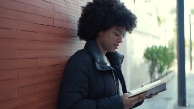 African american woman reading book leaning on wall at street