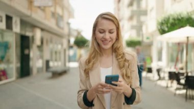 Genç sarışın kadın sokakta akıllı telefon kullanarak gülümsüyor.