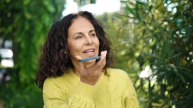 Orta yaşlı İspanyol kadın gülümsüyor. Parkta akıllı telefonla mesaj yolluyor.