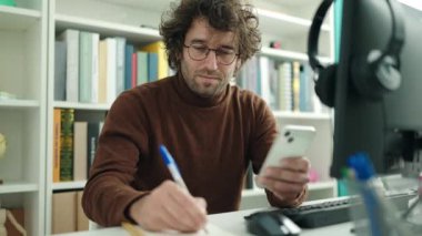 Young hispanic man student using smartphone writing on notebook at library university