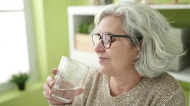 Middle age woman with grey hair drinking glass of water sitting on table at home