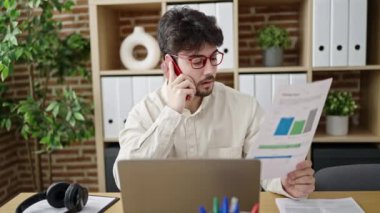 Young hispanic man business worker talking on smartphone reading document at office
