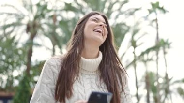 Genç, güzel, İspanyol bir kadın sokakta akıllı telefon kullanarak gülümsüyor.