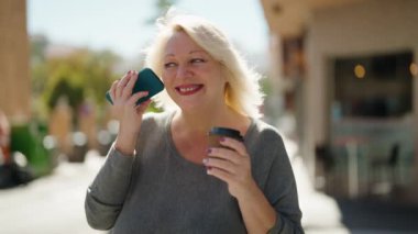 Middle age blonde woman listening audio message by the smartphone drinking coffee at street