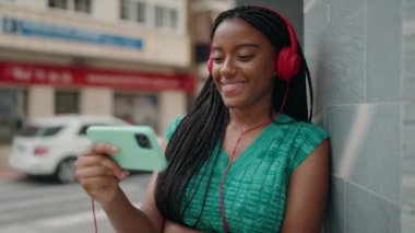 Afro-Amerikalı kadın gülümsüyor. Sokakta akıllı telefondan videolar izliyor.