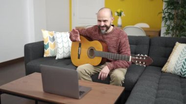 Genç kel adam evdeki kanepede oturmuş online klasik gitar dersi alıyor.