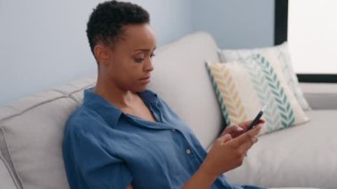 Evdeki koltukta oturan Afro-Amerikan kadın akıllı telefon kullanıyor