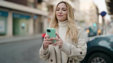 Genç sarışın kadın, kendinden emin gülümsüyor. Sokakta akıllı telefonun yanında özçekim yapıyor.