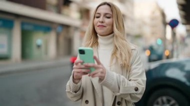 Young blonde woman smiling confident making selfie by the smartphone at street