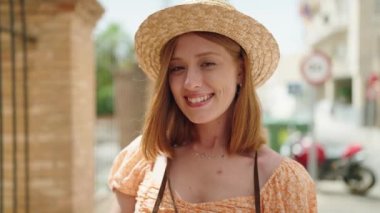 Sokakta klasik kamera kullanan, yaz şapkası takan genç, kızıl saçlı bir kadın.