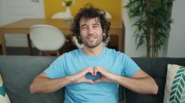 Young hispanic man doing heart gesture over eye sitting on sofa at home