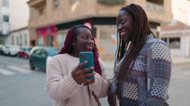 Two african american women smiling confident using smartphone at street