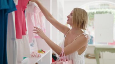 Young blonde woman customer smiling confident holding clothes on rack at clothing store