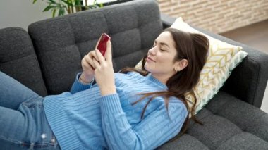 Beyaz kadın akıllı telefon kullanıyor. Evdeki kanepede yatıyor.