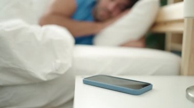 Young hispanic man lying on bed turning off smartphone alarm sleeping at bedroom