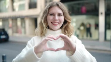 Young blonde woman doing heart gesture with hands over eyes at street