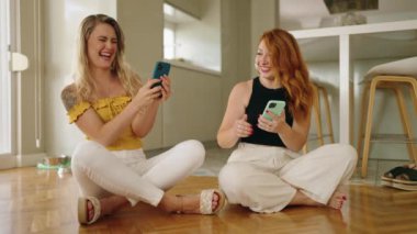 Two women using smartphones sitting on floor at home