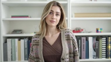 Young blonde woman student standing with confused expression at library university