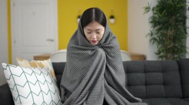 Young chinese woman covering with blanket for cold sitting on sofa at home