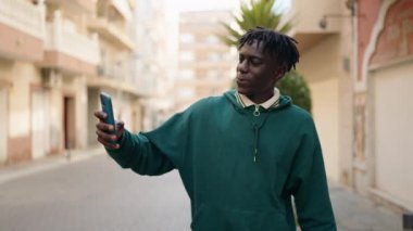 Afro-Amerikalı adam gülümsüyor. Kendine güveni tam. Sokakta akıllı telefondan selfie çekiyor.