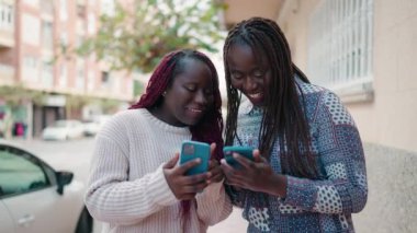 Two african american women smiling confident using smartphone at street