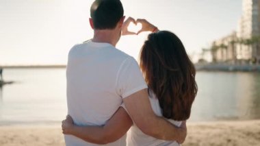 Erkek ve kadın çift birbirlerine sarılıyor. Elleriyle kumsalda kalp hareketi yapıyorlar.