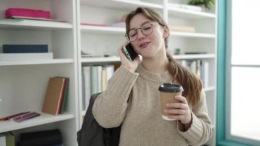 Young blonde woman student talking on smartphone drinking coffee at library university