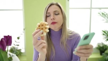 Genç sarışın kadın evde akıllı telefon kullanarak waffle yiyor.